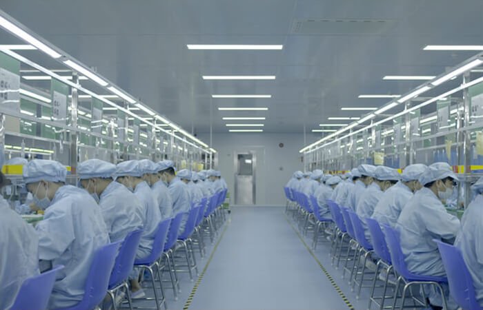 Factory workers wearing protective suits, masks, and hairnets operating on a cleanroom assembly line at the uKERA hardware manufacturing facility.